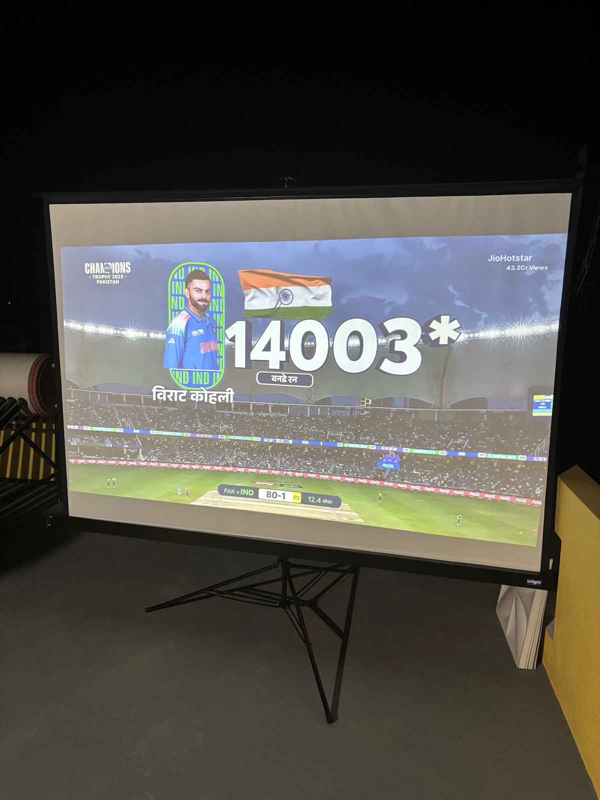 An outdoor projector screen displays a cricket match with illuminated stadium lights and a scoreboard showing a player’s statistics. The Indian flag is prominently featured, alongside the player’s name, creating a lively atmosphere for sports enthusiasts.