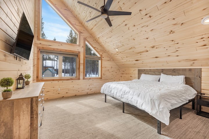A bright, spacious room featuring a king bed with crisp bedding, pillows, a dresser, a mounted TV, and a ceiling fan. Large windows fill the space with natural light and offer peaceful forest views.