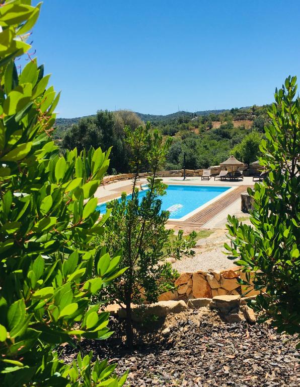 A serene outdoor area is framed by lush greenery, leading to a clear blue swimming pool. Sun loungers are positioned around the pool, with a shaded seating area visible in the background. The landscape features gentle hills, enhancing the tranquil setting.