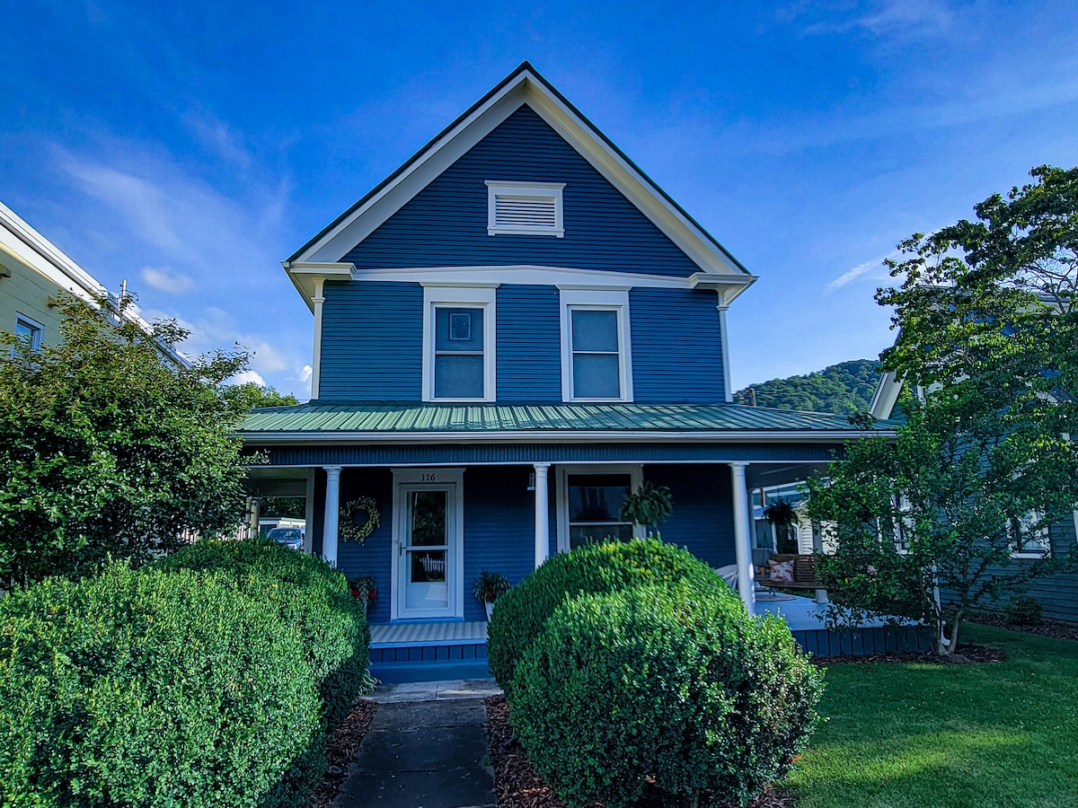The exterior of a charming blue Victorian home is captured under a clear sky. Lush, rounded bushes frame the entrance, and a welcoming porch extends across the front, complemented by a green metal roof and architectural details, inviting guests to relax outside.