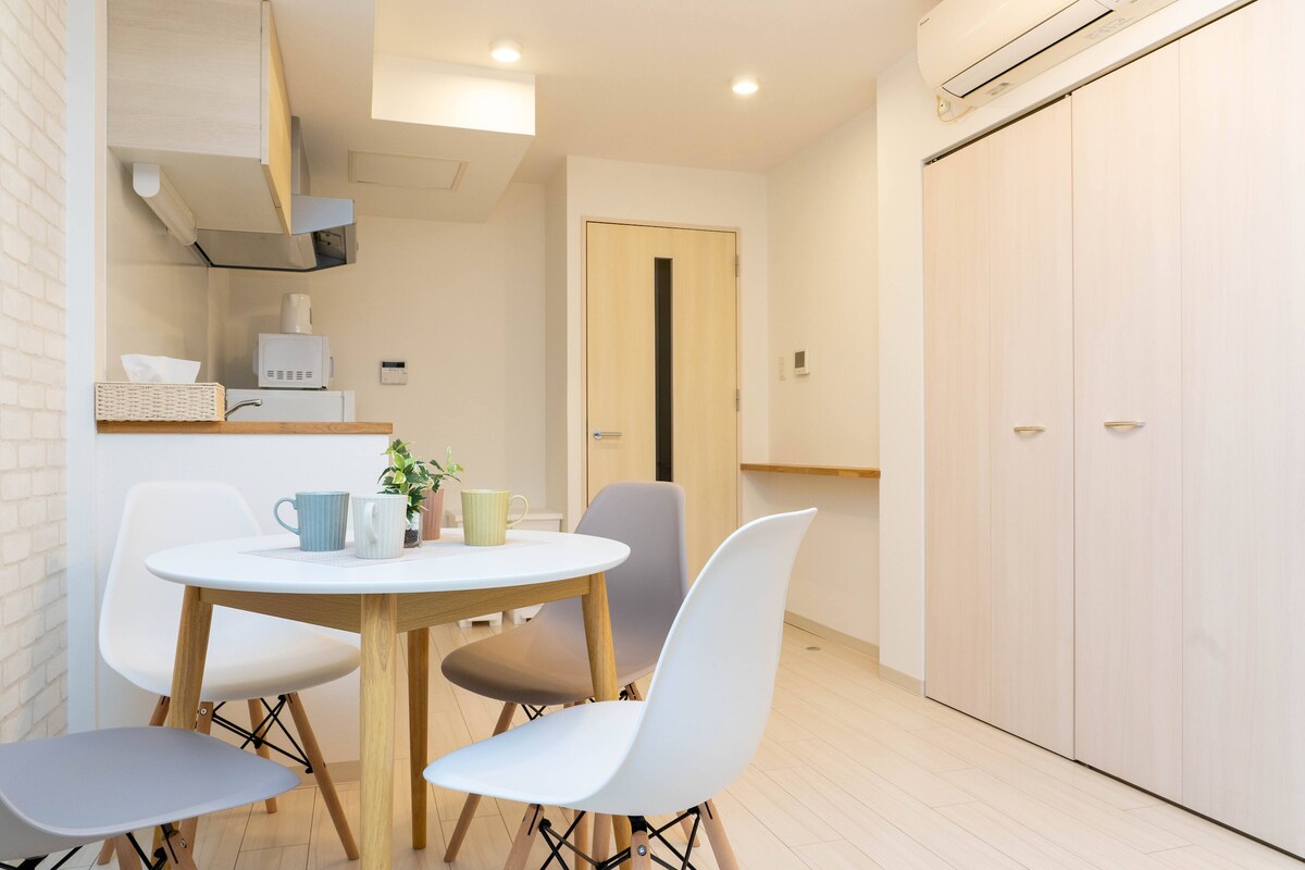 A bright dining area features a round table surrounded by four chairs in varying colors. Light wood accents are present, along with a small plant placed centrally on the table. The space is complemented by white walls and a simple kitchen area in the background.