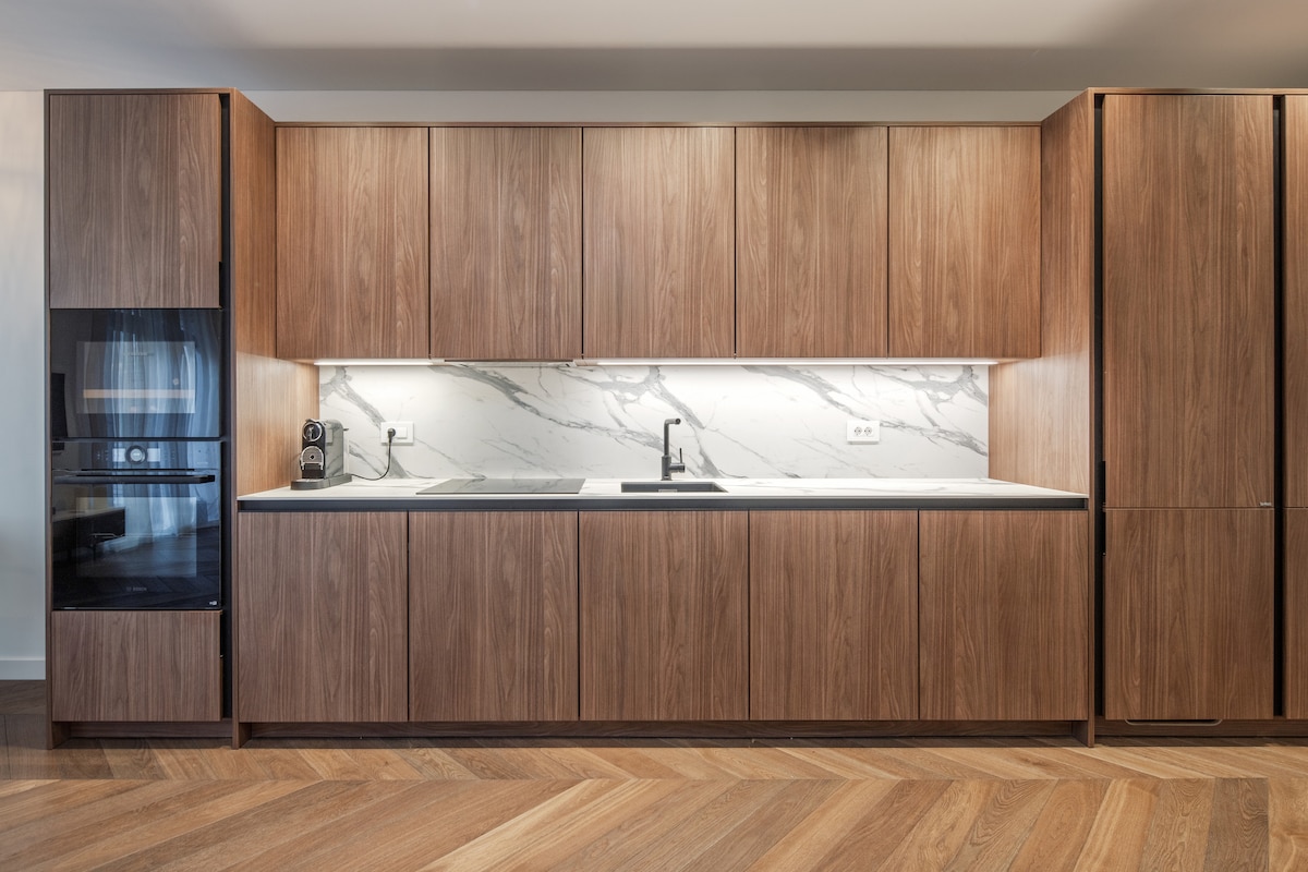 A modern kitchen showcases sleek wooden cabinetry against a marble backsplash, providing a stylish contrast. High-end appliances are integrated into the cabinetry, with a dark sink and faucet centrally positioned on the countertop. The warm wooden flooring complements the contemporary design.