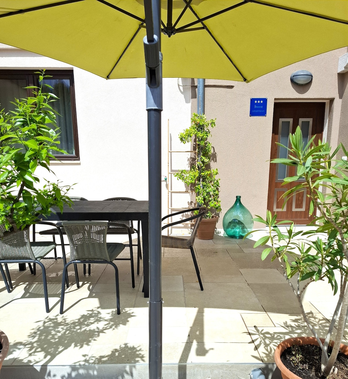 An outdoor terrace area features a large yellow umbrella providing shade over a dining table surrounded by several chairs. Potted plants are arranged around the space, and a decorative glass bottle is placed beside a wooden ladder against the wall.