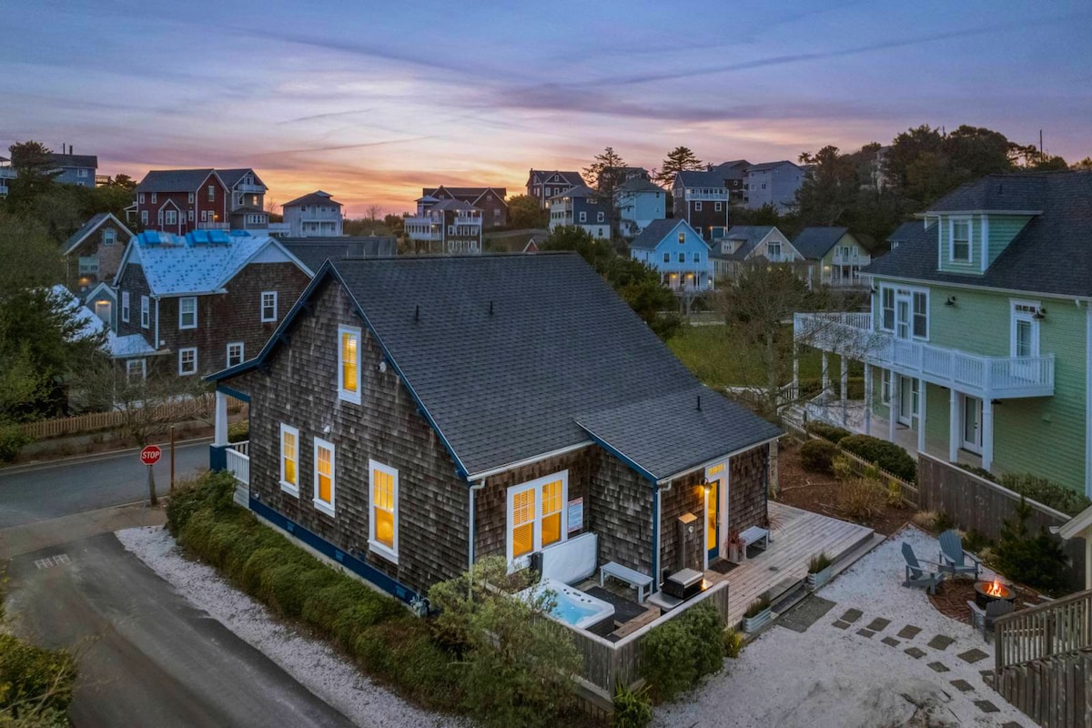 An aerial view showcases the charming Olivia Beach Bungalow's exterior with wooden siding and a sloped roof. The welcoming front porch features outdoor seating and a hot tub, all surrounded by landscaped greenery. Behind, colorful houses of the Olivia Beach community are visible under a serene twilight sky.