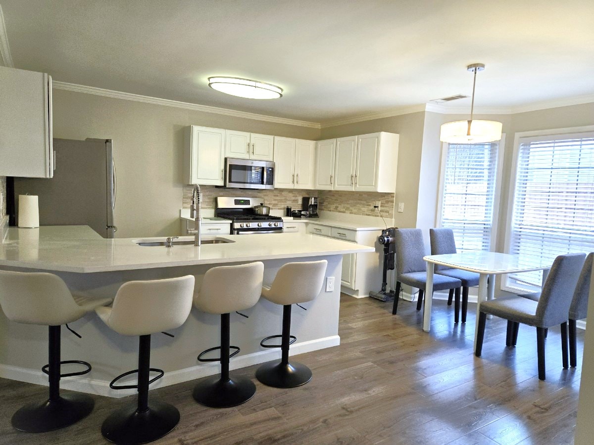 A modern kitchen area featuring sleek white cabinetry and stainless-steel appliances. A spacious island with four high-backed bar stools provides additional seating. A dining table with grey upholstered chairs sits nearby, illuminated by natural light coming through the windows.