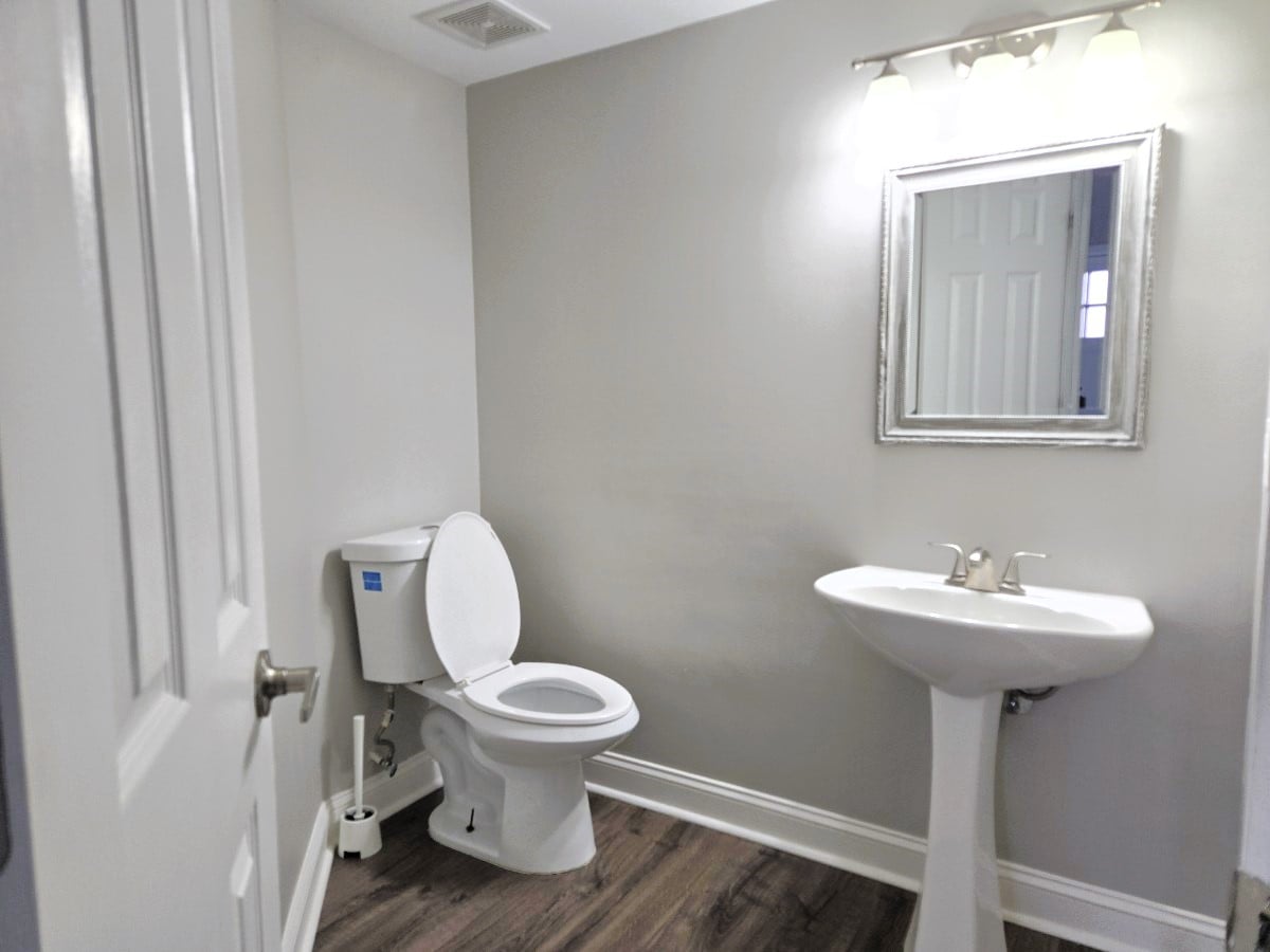 A clean and modern bathroom is presented, featuring a white toilet and a sleek pedestal sink. A decorative mirror is mounted above the sink, and neutral walls complement the wood-style flooring.
