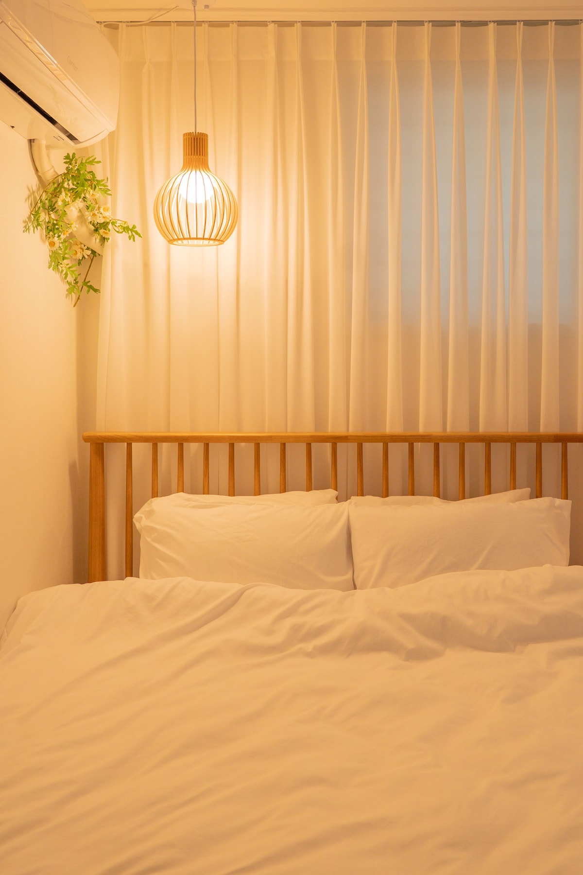 A cozy sleeping area features a spacious bed with white linens. A soft glow from a pendant light above gently illuminates the space. Light-filtering curtains provide privacy while allowing natural light to brighten the room.