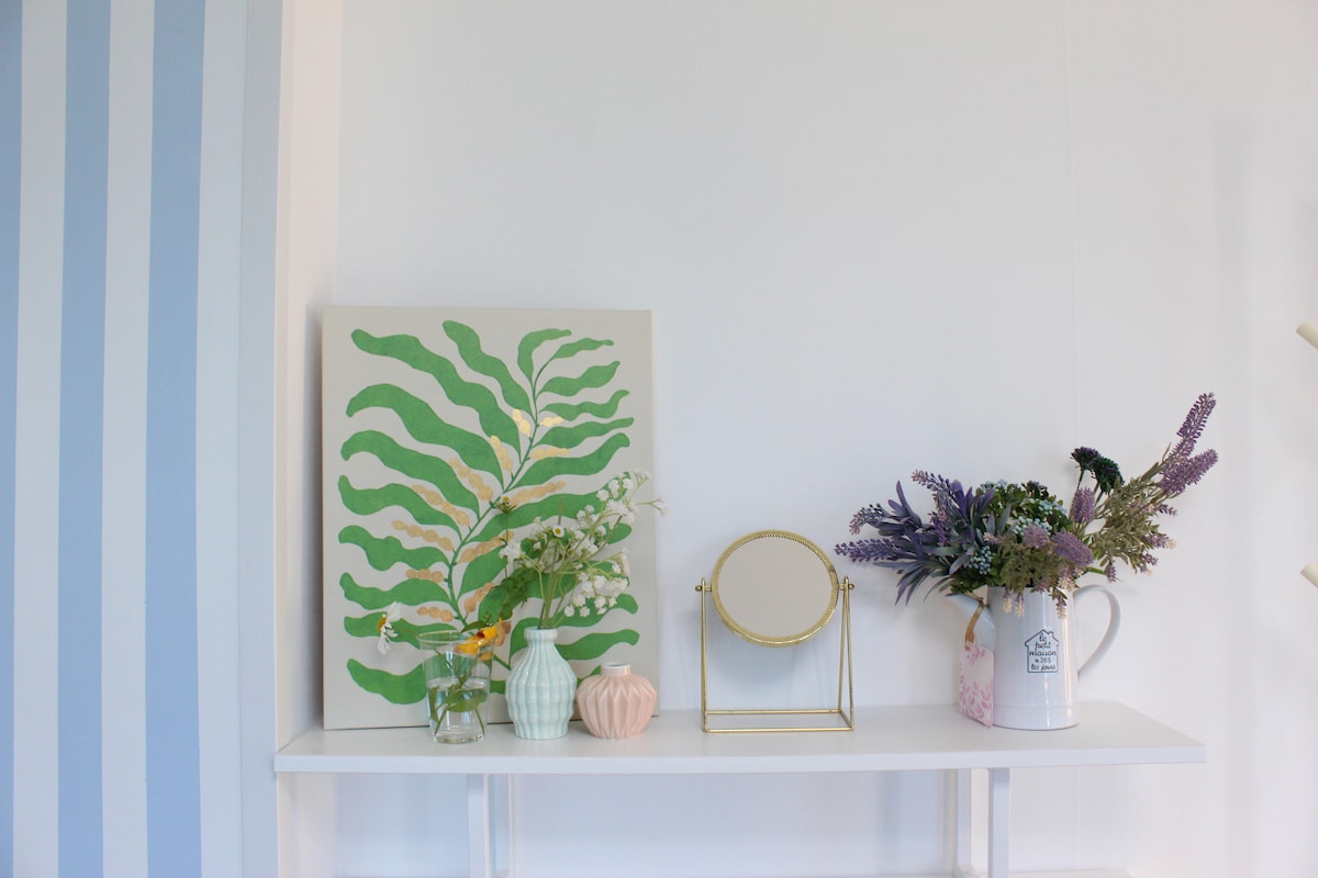 A stylish shelf displays a collection of decorative items, including a vibrant green wall art piece, various vases with flowers, and a small mirror. The arrangement is complemented by a softly colored jug, adding an appealing touch to the space.