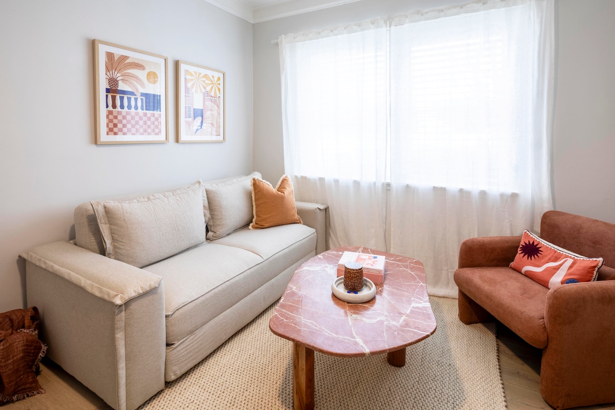 A comfortable seating area is highlighted by a soft, neutral-toned sofa accompanied by an orange accent pillow. A cozy, rust-colored chair complements a round, marble-top coffee table decorated with a bowl and a candle. Natural light filters through sheer curtains at the window.