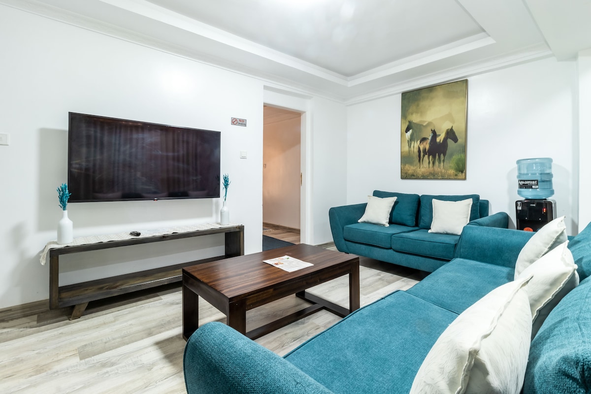 A modern living area features a large flat-screen TV mounted on the wall above a rustic wooden console. Two plush teal sofas are positioned around a dark wooden coffee table, enhancing the space's comfort. A water dispenser and wall art depicting horses add functional and aesthetic elements.