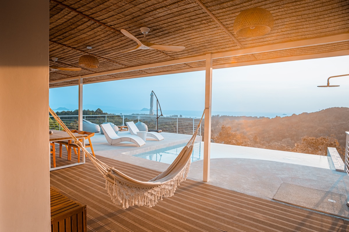 A spacious outdoor terrace features a hammock and stylish sun loungers, offering ocean views. The saltwater pool reflects the sky, while a comfortable dining area is set to the side. Ceiling fans provide airflow under the natural woven canopy, enhancing the serene atmosphere.