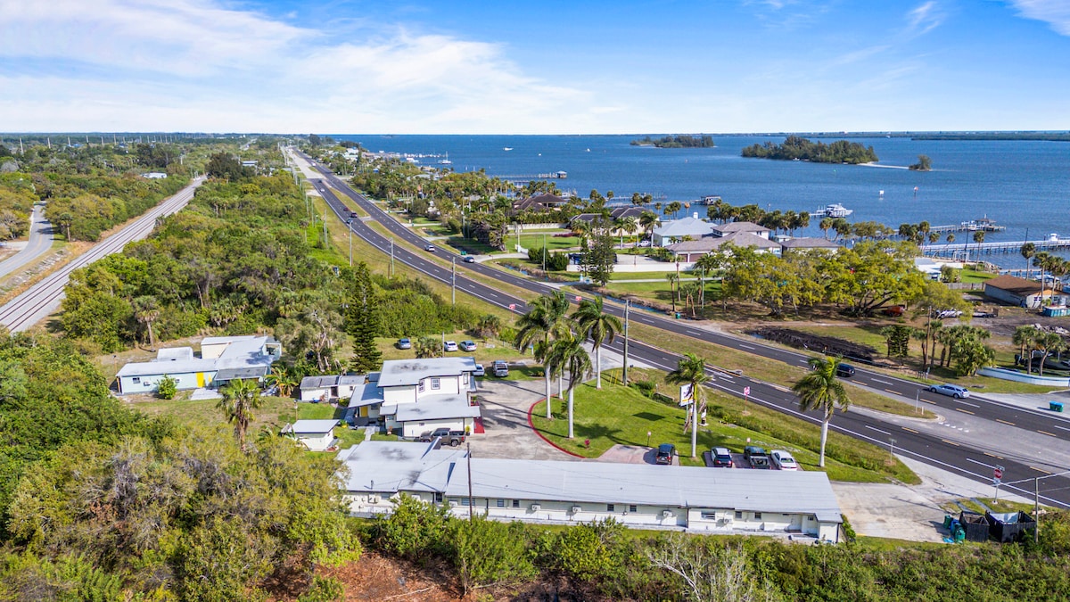 An aerial view highlights the scenic surroundings featuring a blend of greenery and waterways. A coastal road runs parallel to the shoreline, with residential homes and docks visible along the water's edge. Palm trees are scattered throughout the area, enhancing the tropical feel.