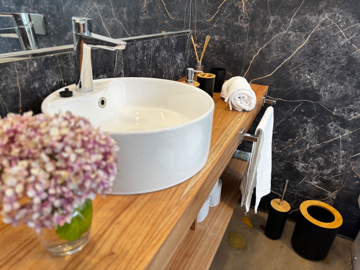 A modern bathroom features a round white sink atop a wooden vanity, complemented by a marble backdrop. Fresh flowers are placed in a small glass vase. Towels are neatly hung on a rack, and black and wooden containers are positioned for storage.