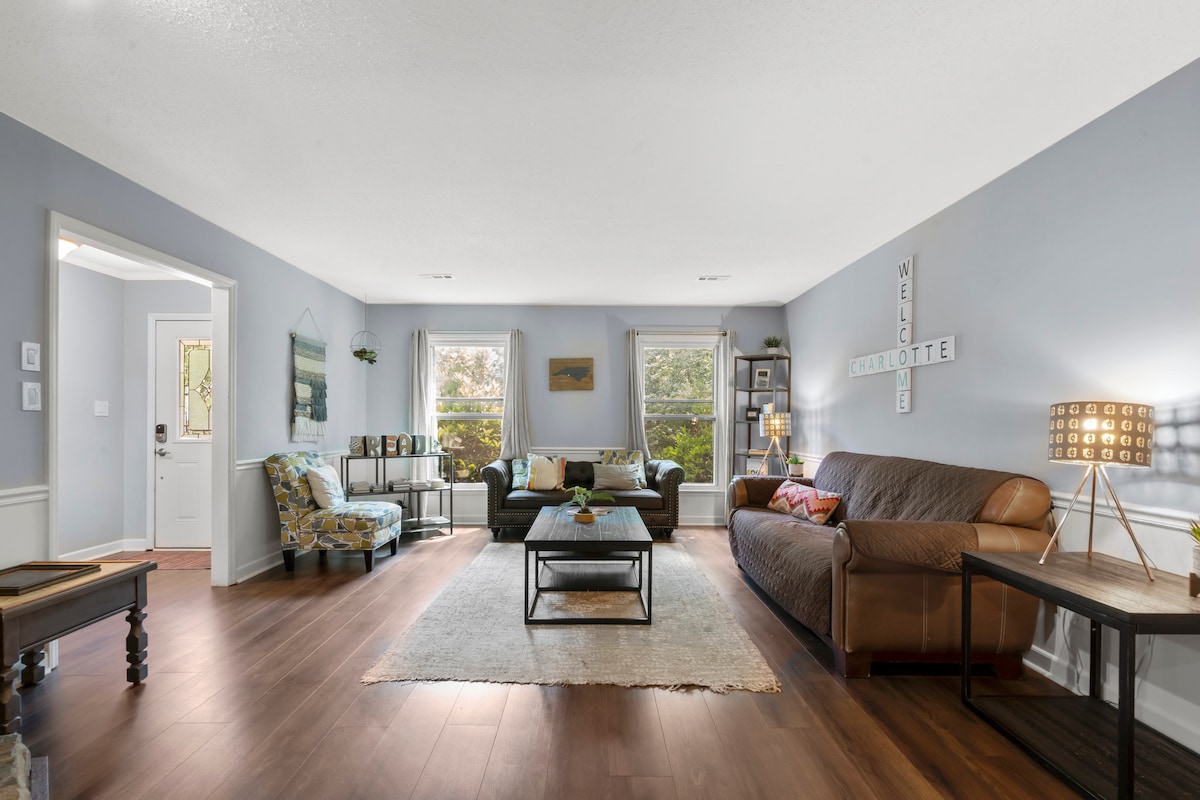 A spacious living area is furnished with a mix of cozy seating options, including a leather sofa and patterned chairs. Natural light filters through the windows, enhancing the neutral wall tones. A simple coffee table centers the room, complementing a light area rug.