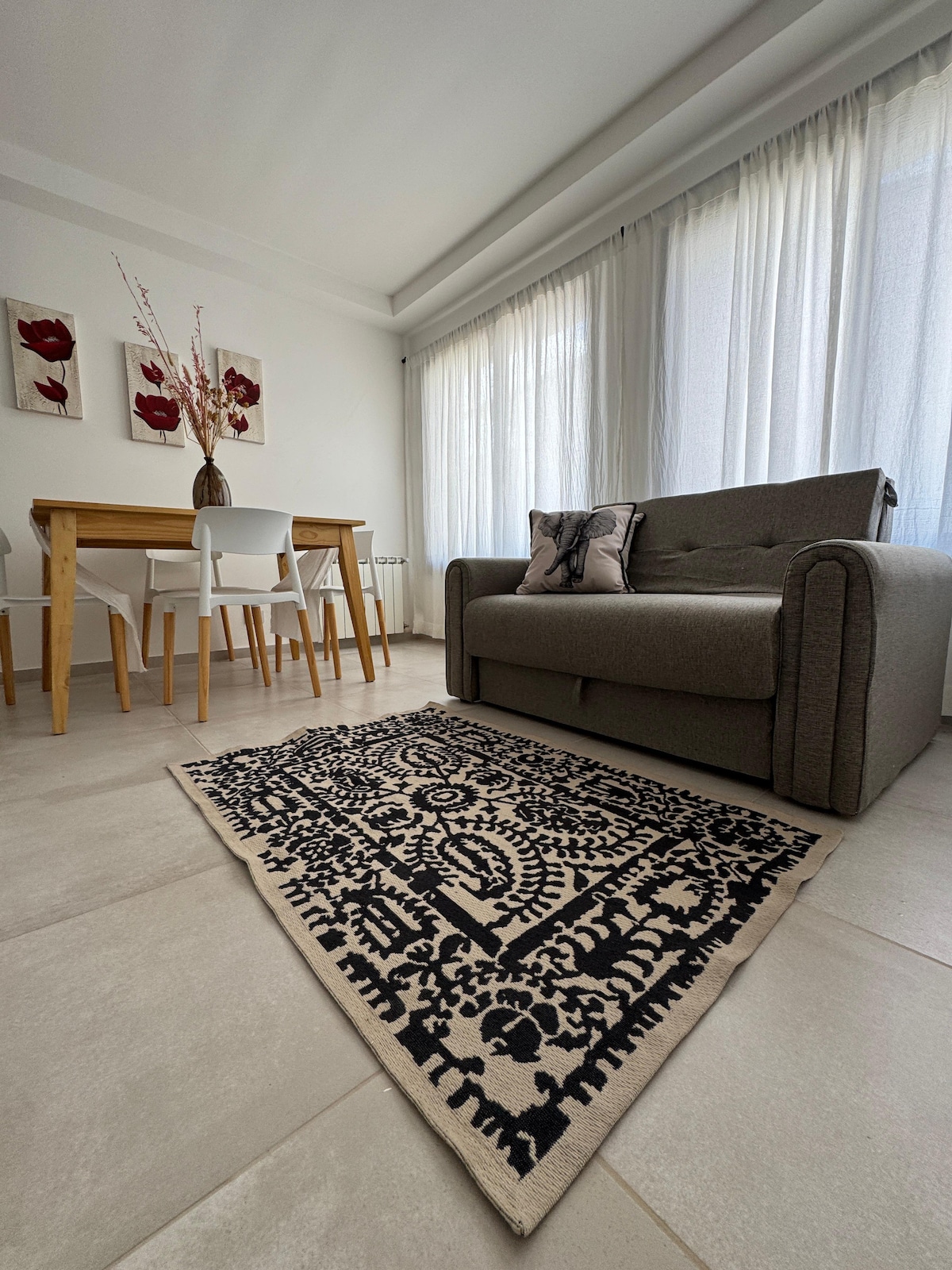 A light-filled living space features a gray sofa facing a dining table surrounded by white chairs. A decorative area rug with intricate patterns is placed on the tiled floor, enhancing the room's inviting ambiance. Large windows allow natural light to illuminate the space.