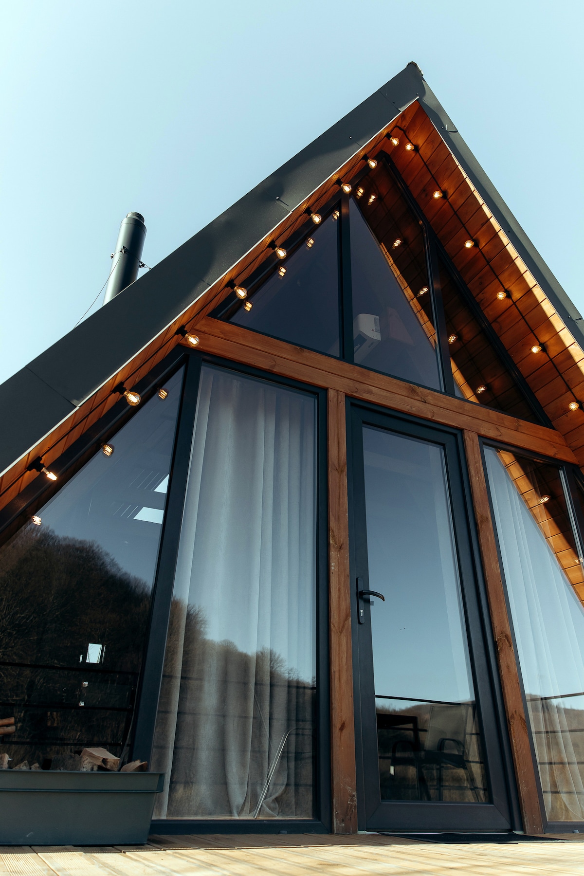 The exterior of a triangular-shaped cabin is highlighted with warm string lights. Large glass doors offer a view into the interior, framed by wooden accents and white drapes. The clean lines of the architecture create a modern and inviting presence against a clear blue sky.