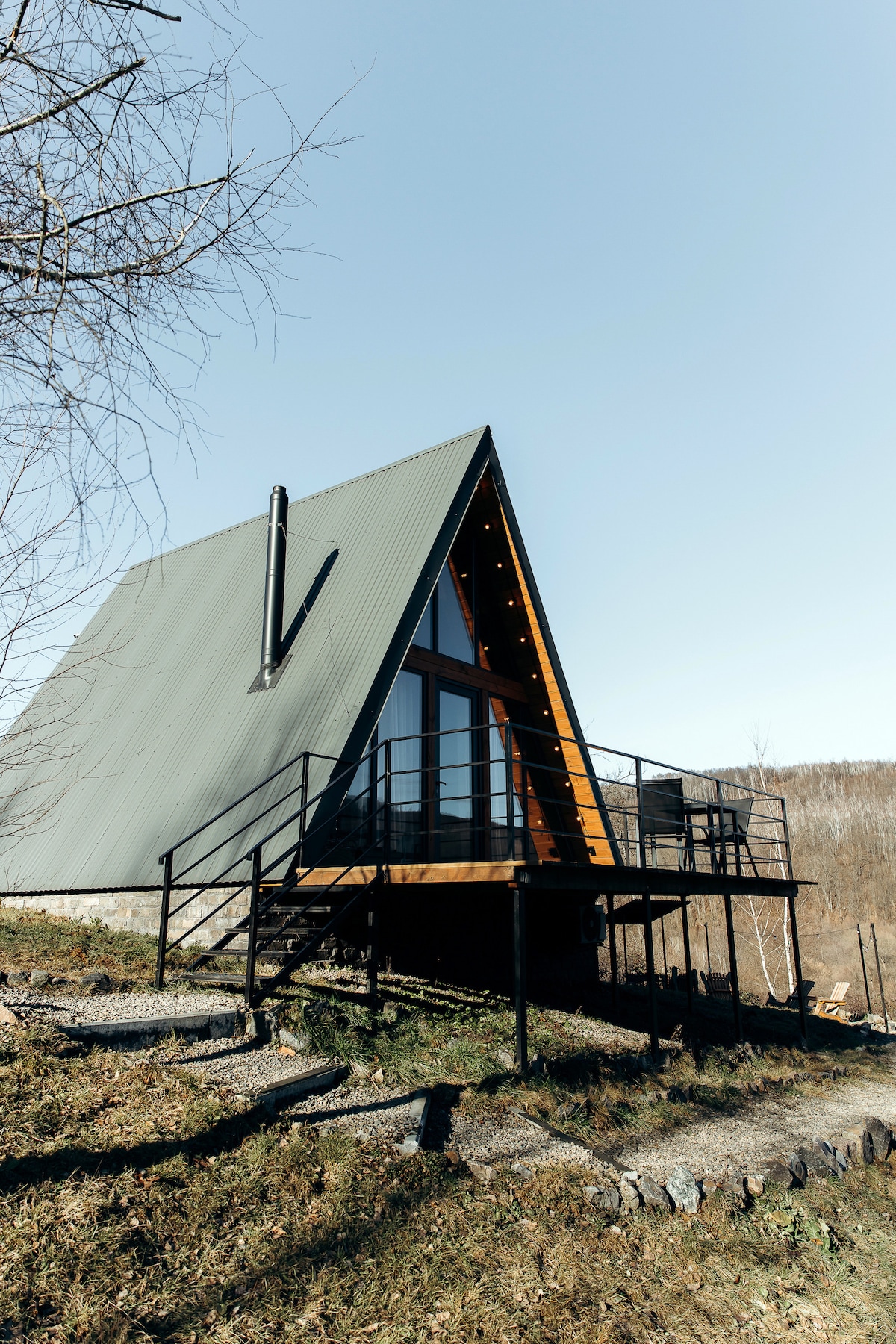 A triangular cabin with large glass windows and a sloped roof is set against a clear blue sky. The entrance features a wooden deck with black railings, surrounded by grass and sparse trees. Scenic views of the surrounding nature can be appreciated.