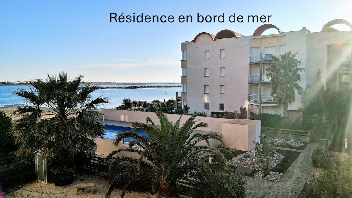 A seaside residence is showcased against a clear sky, featuring a landscaped garden and an outdoor pool. Palm trees and stone pathways lead toward the exterior of the building, enhancing the coastal ambiance and providing glimpses of the water from the grounds.