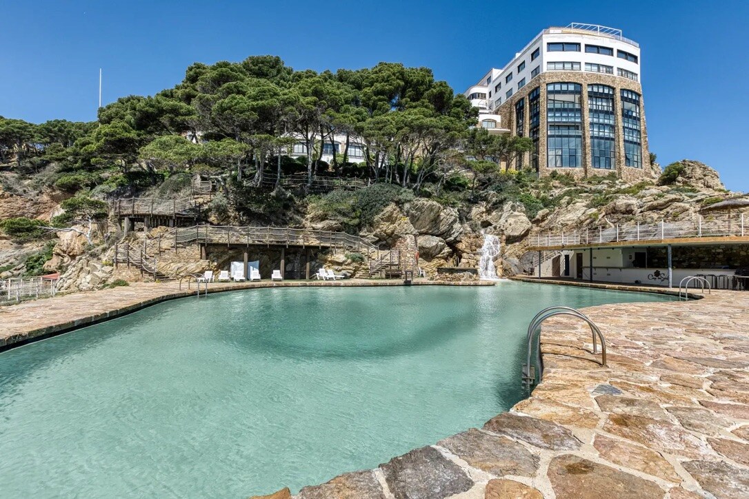 An outdoor pool is framed by natural stone and surrounded by landscaped greenery. A modern building with large glass windows overlooks the serene water. Sunlight reflects off the pool's surface, creating a calm atmosphere.