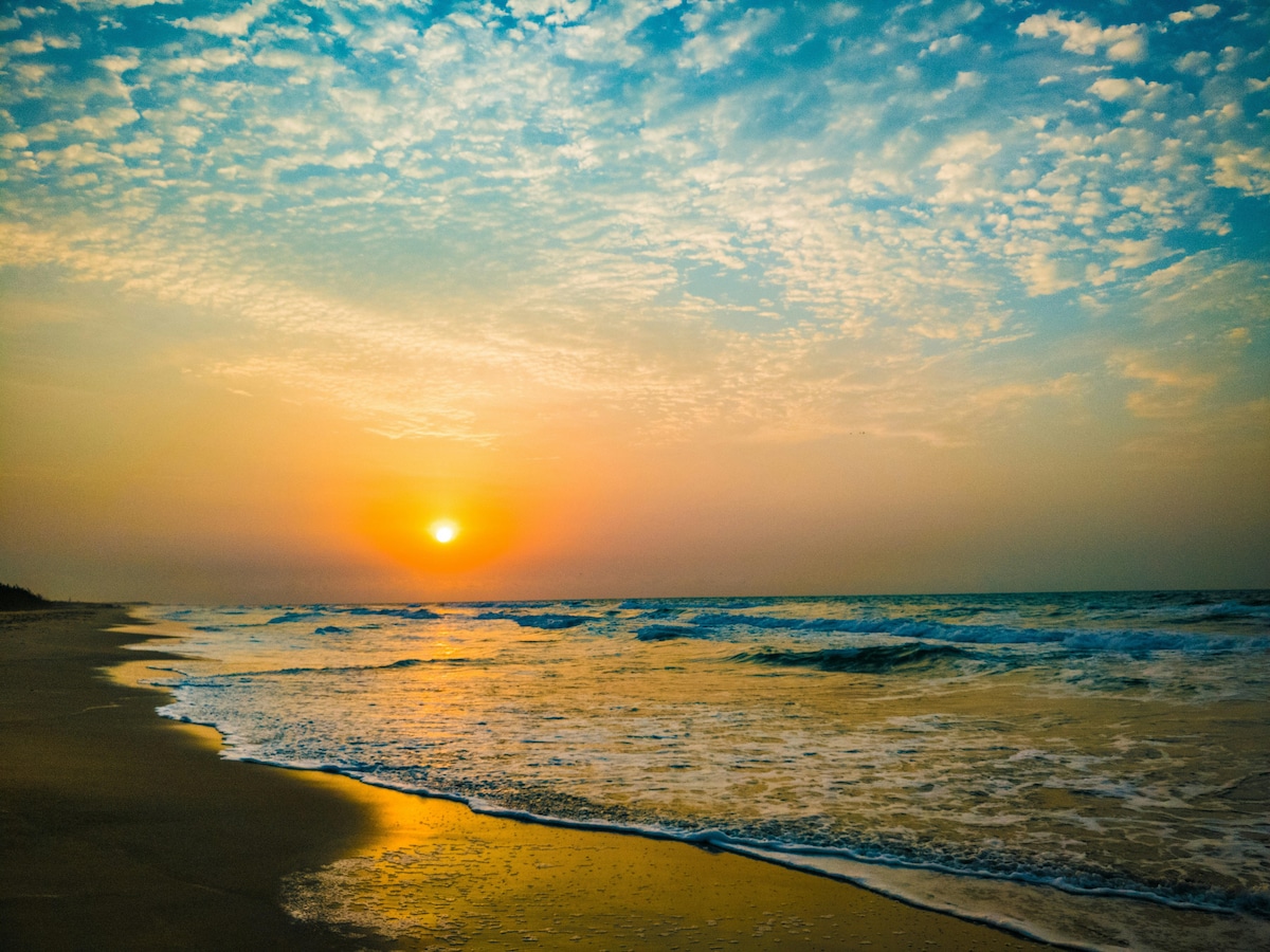 A serene beach scene captures the sun setting on the horizon, casting a warm orange and golden glow across the sky. Gentle waves reflect the colors of the sky, while the sand glistens with the soft light, creating a peaceful coastal atmosphere.