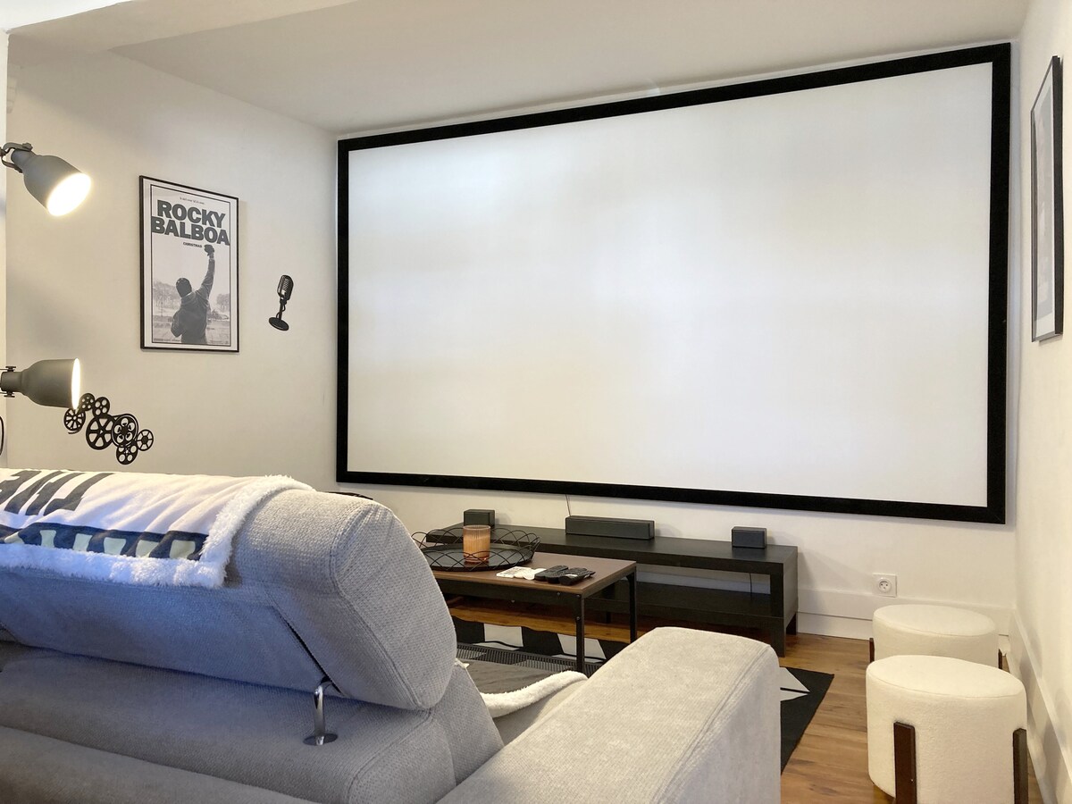 A cozy media area is showcased with a large projection screen framed by a minimalistic black border. A comfortable gray sofa is positioned nearby, accompanied by a coffee table and decorative elements, creating a relaxed environment for movie viewing.
