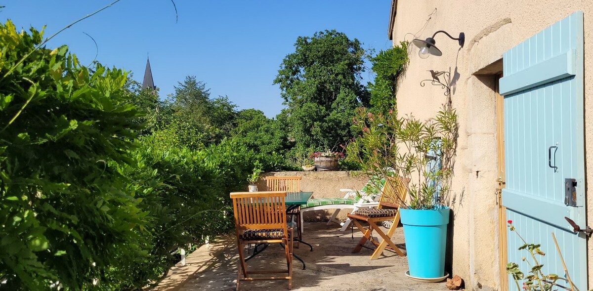 A sunny outdoor terrace features a dining table and four wooden chairs surrounded by lush greenery. A large potted plant adds color beside the blue door, while distant trees and a church steeple are visible in the background, creating a serene setting.