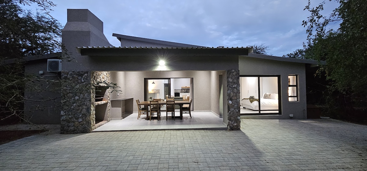 The exterior of the villa features a stone-accented façade and a spacious terrace. Large glass doors open to reveal a dining area with a wooden table and chairs, while a cozy fireplace is visible for outdoor gatherings. Soft light enhances the inviting atmosphere.