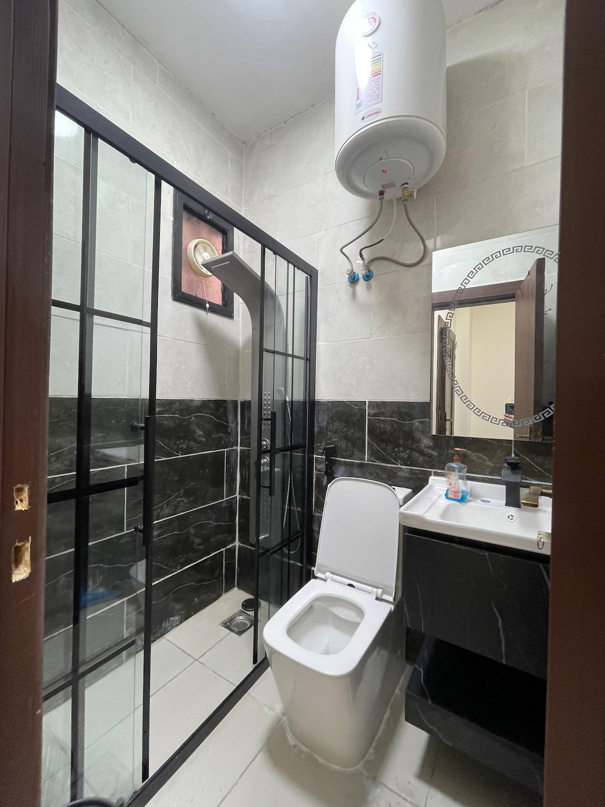 A modern bathroom featuring a glass-enclosed shower with a rainfall showerhead. A sleek black and white color scheme is apparent, with black accents on the wall and a white toilet. A vanity with a mirror and a sink sits adjacent, while a water heater is mounted above.