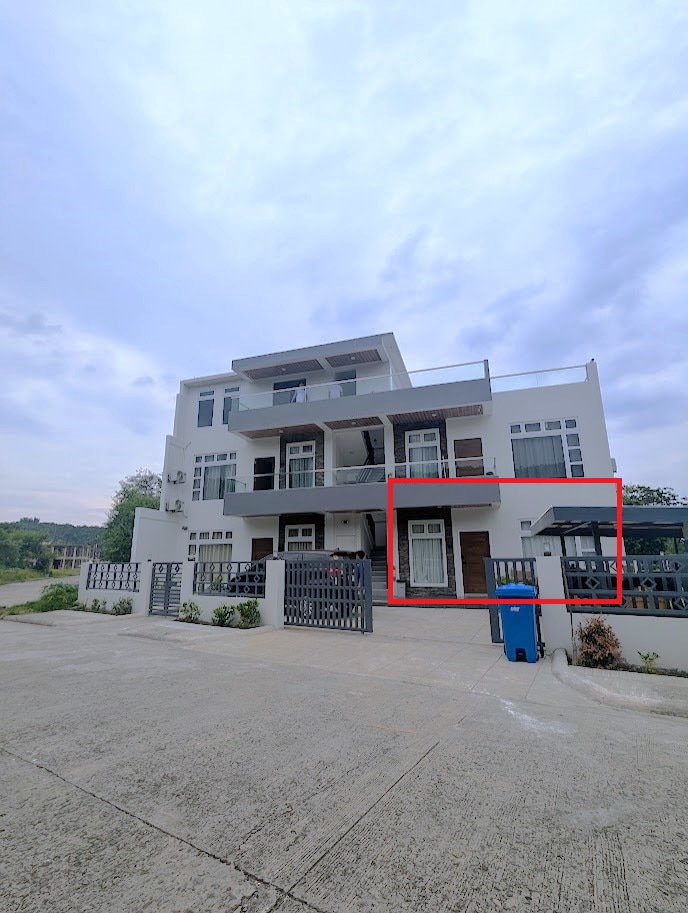 The exterior of a modern three-story condo complex is presented, showcasing a blend of white and grey tones. A balcony is visible on the second floor, framed by glass doors. A secured entrance with a gate is seen in the foreground, leading to the building.