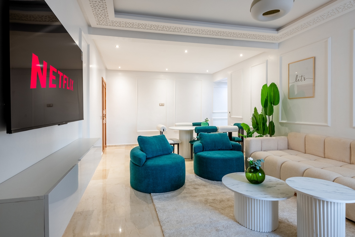 A spacious living area is presented with two teal sofas and a light-colored sectional sofa. A large TV screen is mounted on the wall, displaying the Netflix logo. Natural light fills the room, enhancing the modern decor.
