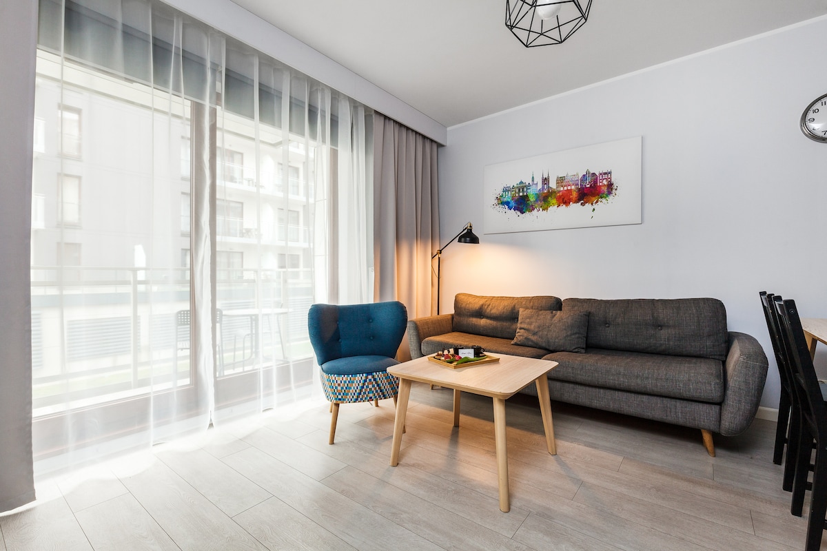 A modern living area is presented with a gray couch and a blue accent chair. Natural light filters through sheer curtains, highlighting a wooden coffee table and a wall art piece featuring colorful cityscape imagery. A dining table is placed nearby, completing the space.