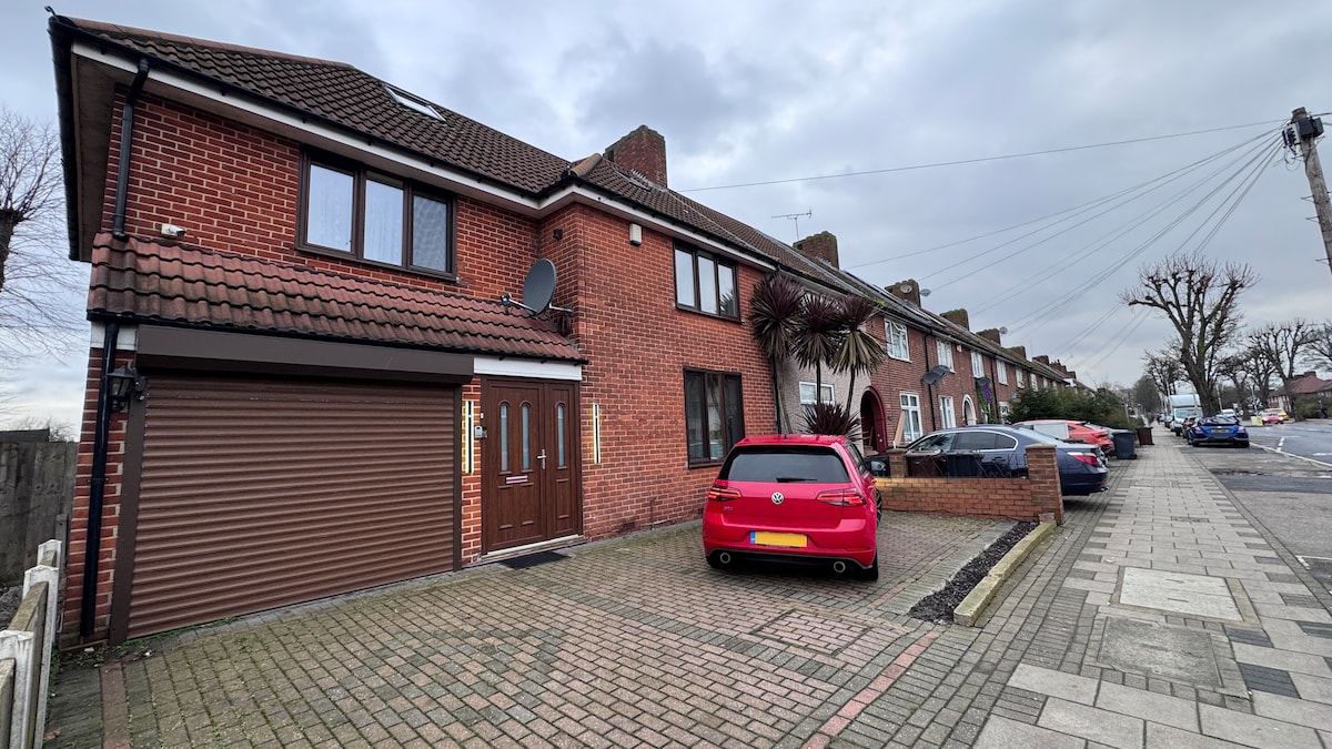 A two-story brick building is shown, featuring a double garage on the left and a driveway accommodating a red vehicle. The front entrance includes two doors, framed by decorative plants. The property is situated in a residential area with a wide sidewalk and adjacent homes.