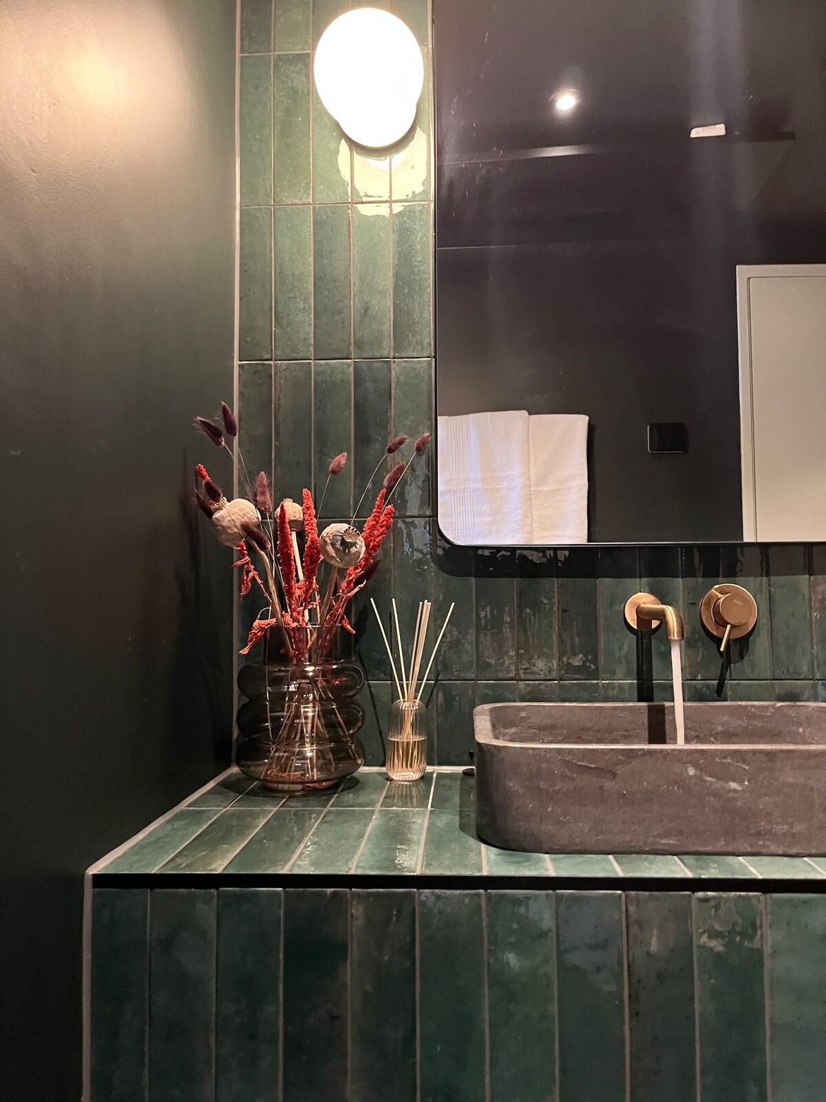 A modern bathroom is depicted with dark green tiles covering the walls. A sleek, stone sink is situated on a countertop, accompanied by a decorative vase of dried flowers. A large mirror hangs above the sink, reflecting the elegant lighting and providing a sense of depth.