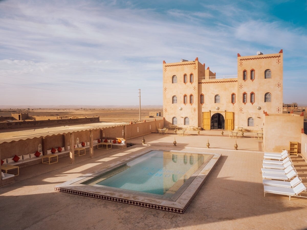 An outdoor area is featured, showcasing a refreshing pool surrounded by lounge chairs. The riad's traditional architecture is visible in the background, accented by arched windows and warm, earthy tones. Expansive desert landscapes stretch out beyond the pool area under a clear blue sky.