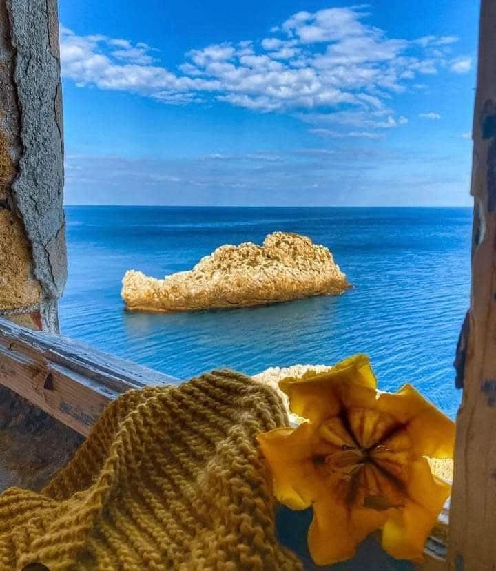 A tranquil view from a window highlights the calm sea, featuring a distant rocky island and a clear blue sky. A soft, knitted blanket and a yellow flower are positioned in the foreground, adding a touch of warmth to the serene landscape.