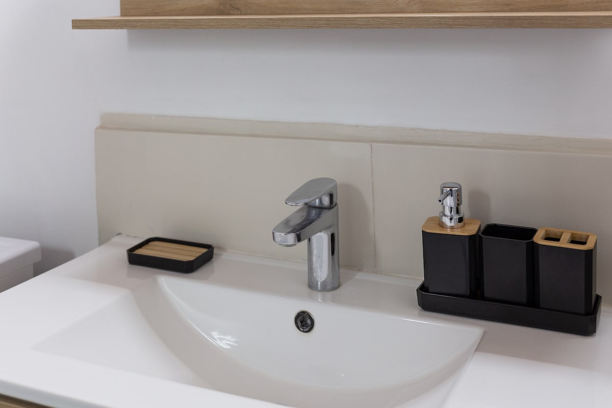 A clean and modern bathroom sink features a sleek faucet above a white basin. To the right, a set of black and wooden containers holds toiletries, while a wooden tray is placed to the left of the sink.