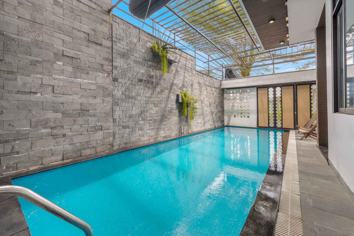 An indoor pool features a clear blue surface, surrounded by sleek stone walls and greenery. Sunlight filters through a slatted roof, creating a warm ambiance. The pool area is complemented by sliding glass doors, opening to additional outdoor space.