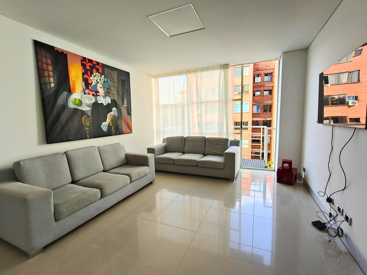 A spacious living area features two light gray sofas arranged for comfort, complemented by large windows allowing natural light to fill the space. A colorful artwork adorns one wall, and a mounted television is positioned for viewing. A balcony is visible through the glass doors.