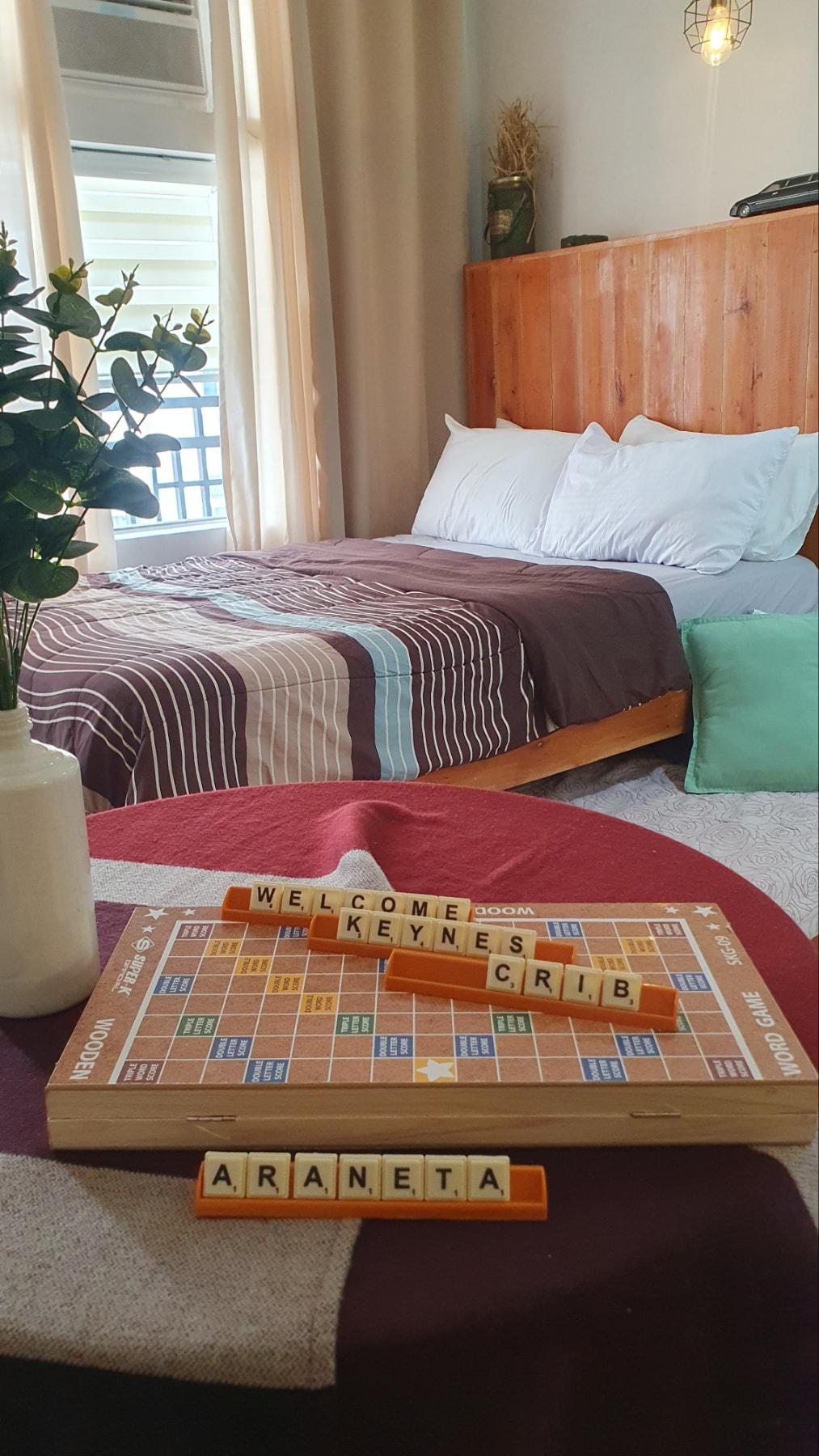 A welcoming studio space is featured, with a double bed in earthy tones and crisp linens. A Scrabble board placed on a small table highlights a leisurely atmosphere. Natural light filters through large windows, complementing the wooden accents and soft furnishings in the room.