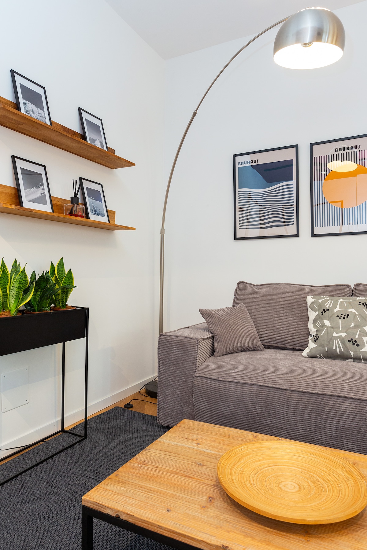 The living area features a comfortable gray sofa adorned with a patterned cushion. A wooden coffee table is centered in front, accompanied by a sleek black side table topped with green plants. Artwork hangs on the wall above floating shelves, enhancing the contemporary decor.