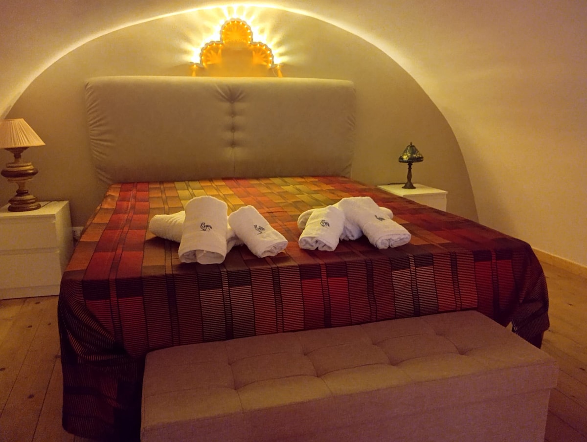 A softly lit sleeping area features a spacious bed adorned with a patterned blanket, complemented by decorative pillows. Fluffy white towels are neatly arranged at the foot of the bed. A lamp and a small decorative table add subtle elements of comfort to the serene atmosphere.