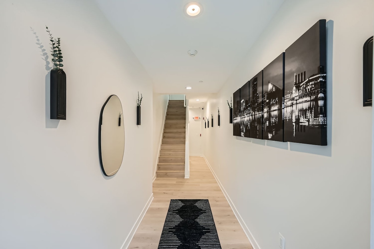 A well-lit hallway features contemporary black and white wall art, adding visual interest to the space. A round mirror is mounted on one wall, and small decorative elements are placed in niches. Soft wood flooring leads to an open staircase at the end of the corridor.