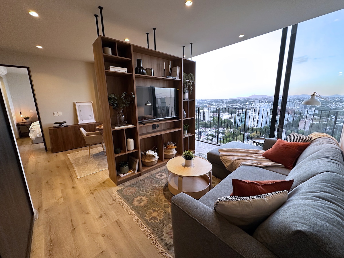 A bright living area features a modern gray sectional sofa adorned with colorful cushions. A light wooden coffee table sits in front, complemented by a patterned area rug. A sleek entertainment unit is set against the wall, providing a panoramic view of the city through expansive glass windows.