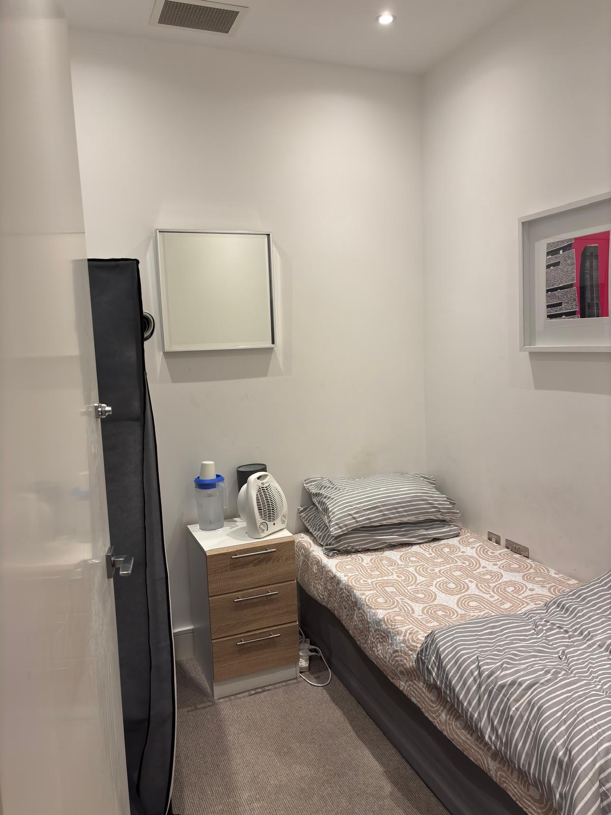 A compact sleeping area features a single bed with striped linens, positioned against a light wall. A bedside table holds a kettle and a lamp. A mirror hangs above the table, while framed art decorates the wall, adding a modern touch to the space.