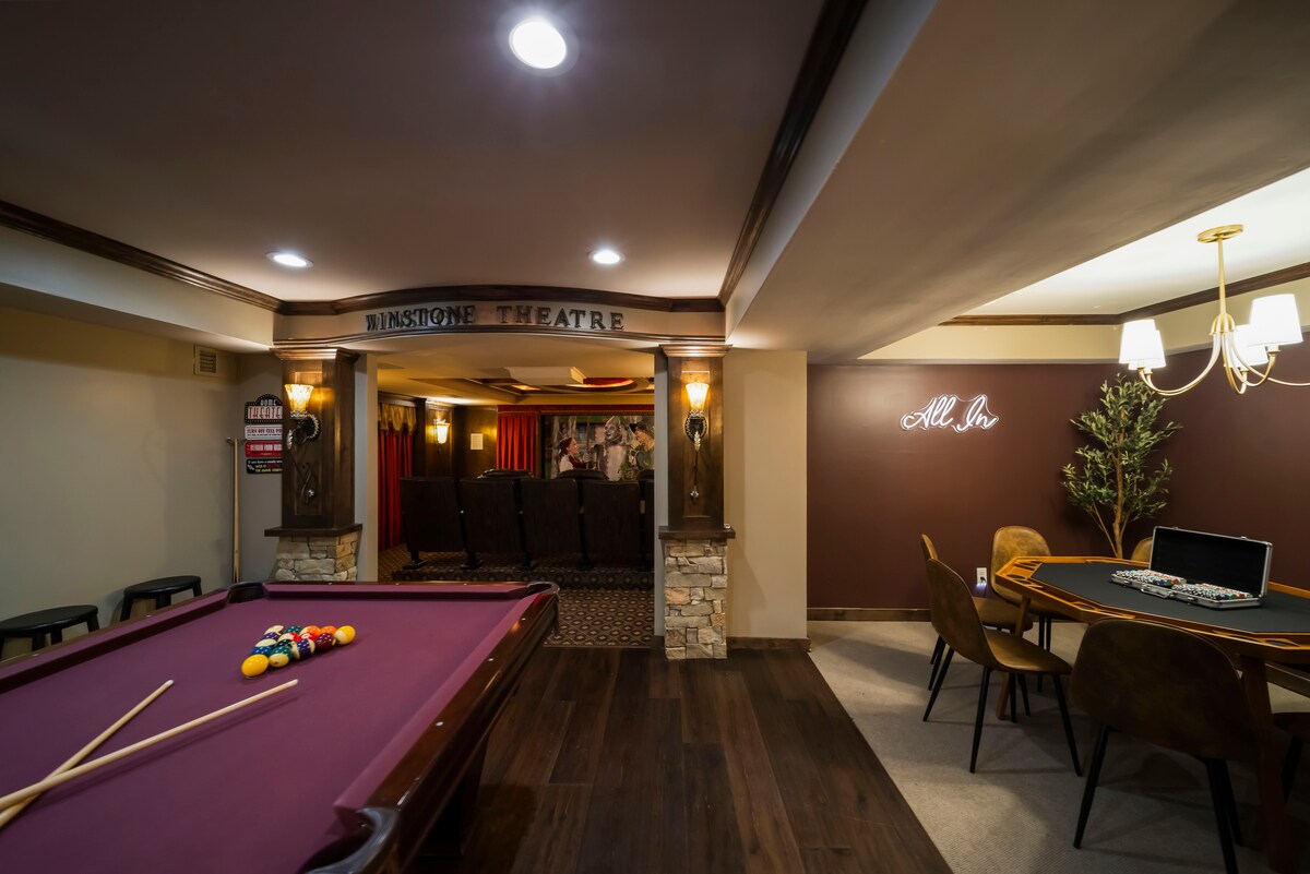 A dedicated gaming area features a billiards table with cues resting on the side, complemented by soft lighting and warm wood accents. The nearby poker area showcases a large table with seating for eight, while a private theater is visible in the background, equipped with rows of spacious seats.