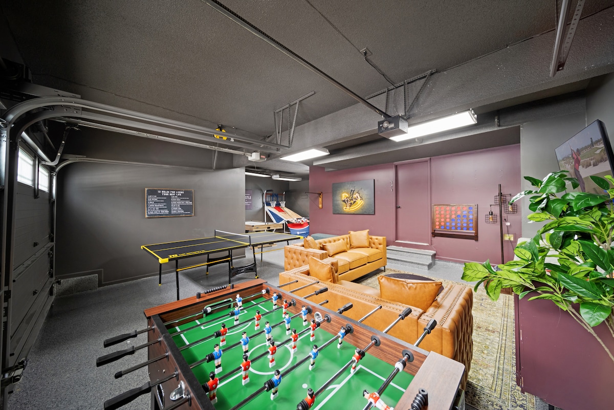 A spacious game room features a green foosball table, air hockey table, and lounge area with leather couches. The space is illuminated by overhead lighting, and artwork adorns the walls. A potted plant adds a touch of greenery to the environment.