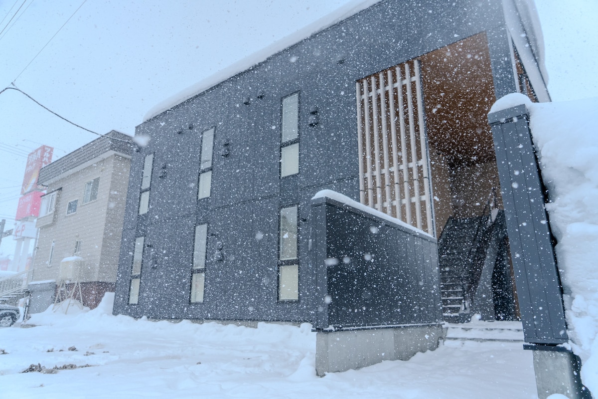 A modern building is presented in a snowy landscape, with large windows and a distinctive vertical wooden slat feature. Snowflakes fall gently, creating a serene winter atmosphere. The entrance is framed by a staircase, leading to the interior of the property.