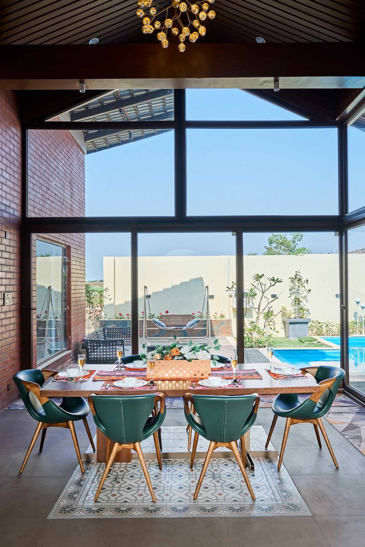 A dining area with a wooden table set for six includes green chairs and decorative plates. Large glass windows offer a view of the outdoor pool and garden, allowing natural light to fill the space. A modern chandelier hangs overhead.