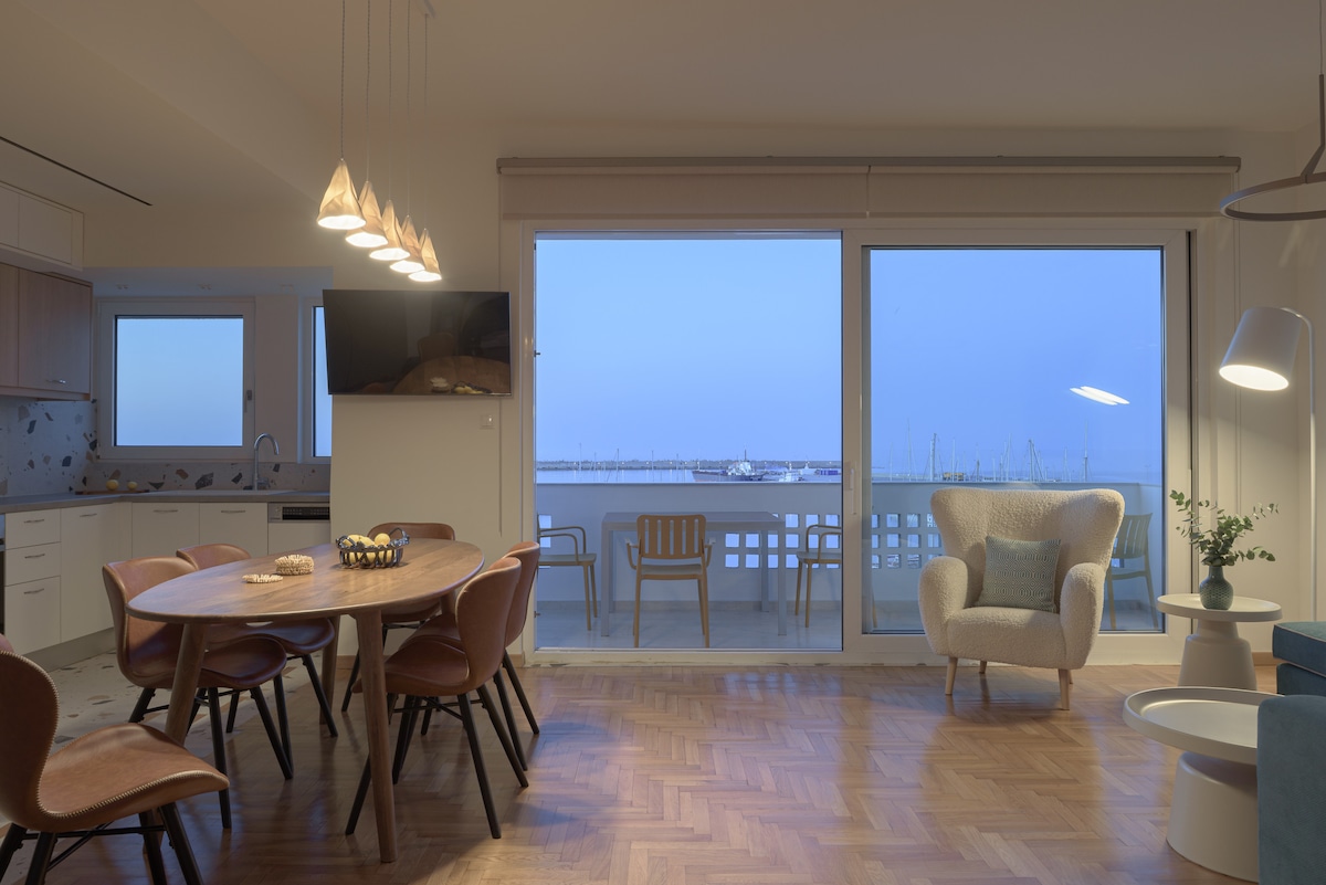A spacious living area is highlighted by large glass doors that offer panoramic views of the sea. A wooden dining table with stylish chairs is paired with a cozy armchair, while soft lighting creates a warm ambiance. The kitchen is visible in the background.