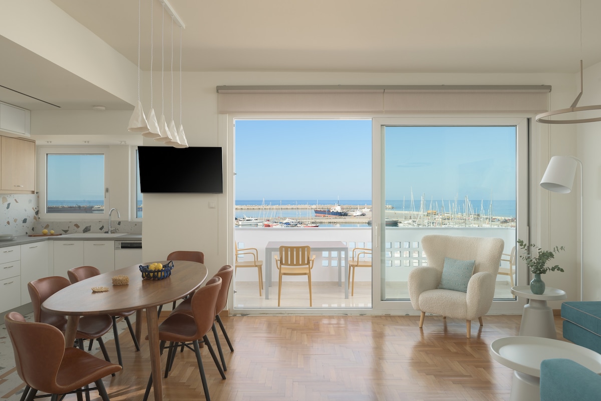 A bright dining area features a round wooden table surrounded by several chairs. A spacious window offers stunning views of the marina and sea, with a cozy armchair placed nearby. Natural light fills the room, highlighting the modern design and furnishings.